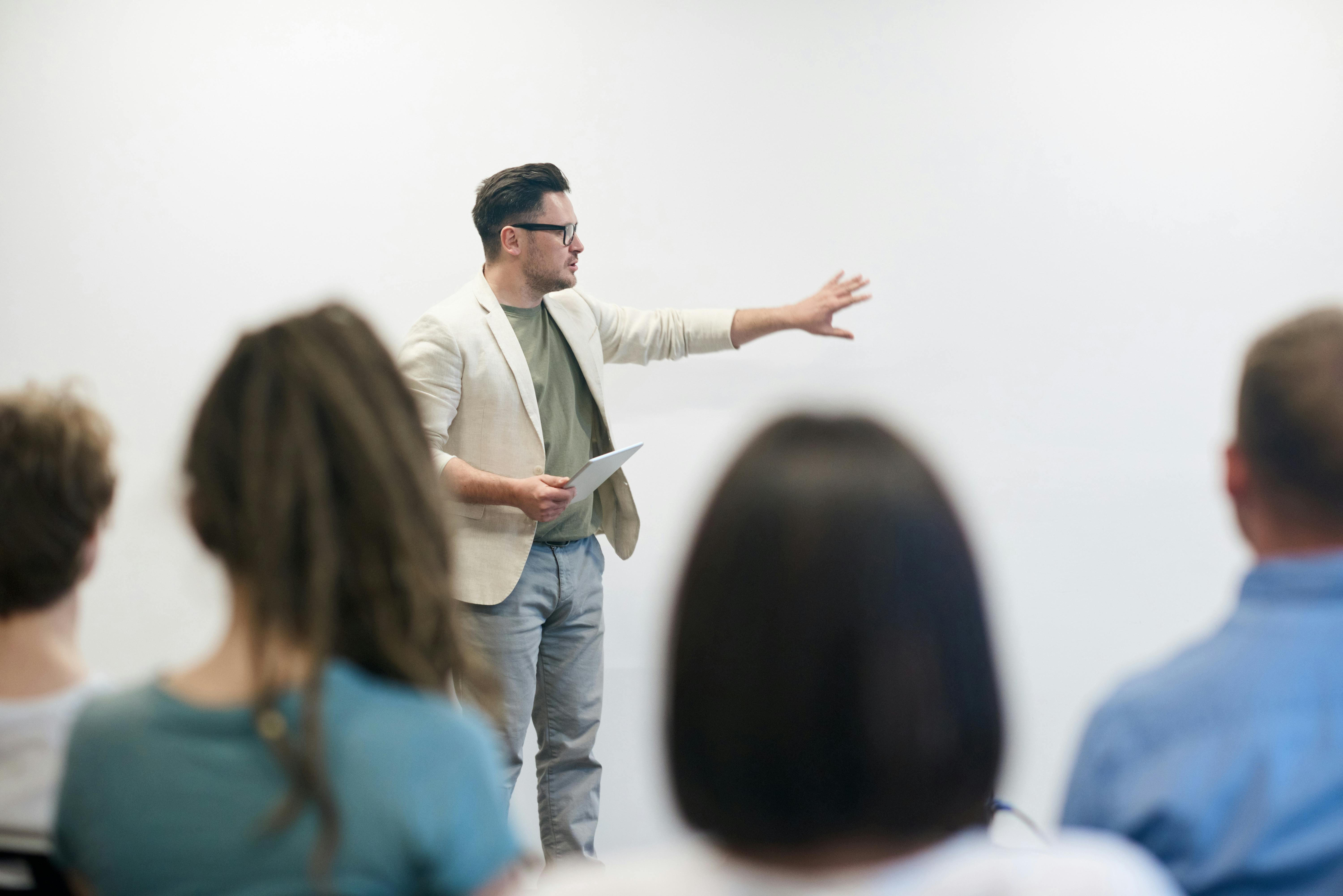  Professor ou palestrante explicando um conceito para um grupo de alunos ou profissionais durante uma apresentação ou aula, com gestos dinâmicos e expressão envolvente.